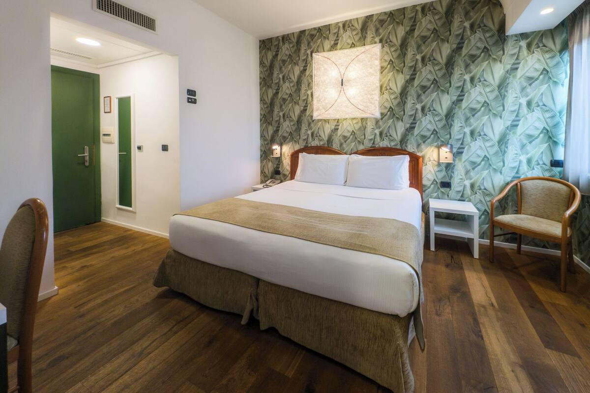 A hotel room with a large bed, a nightstand, a chair, a green door, and a patterned wallpaper.