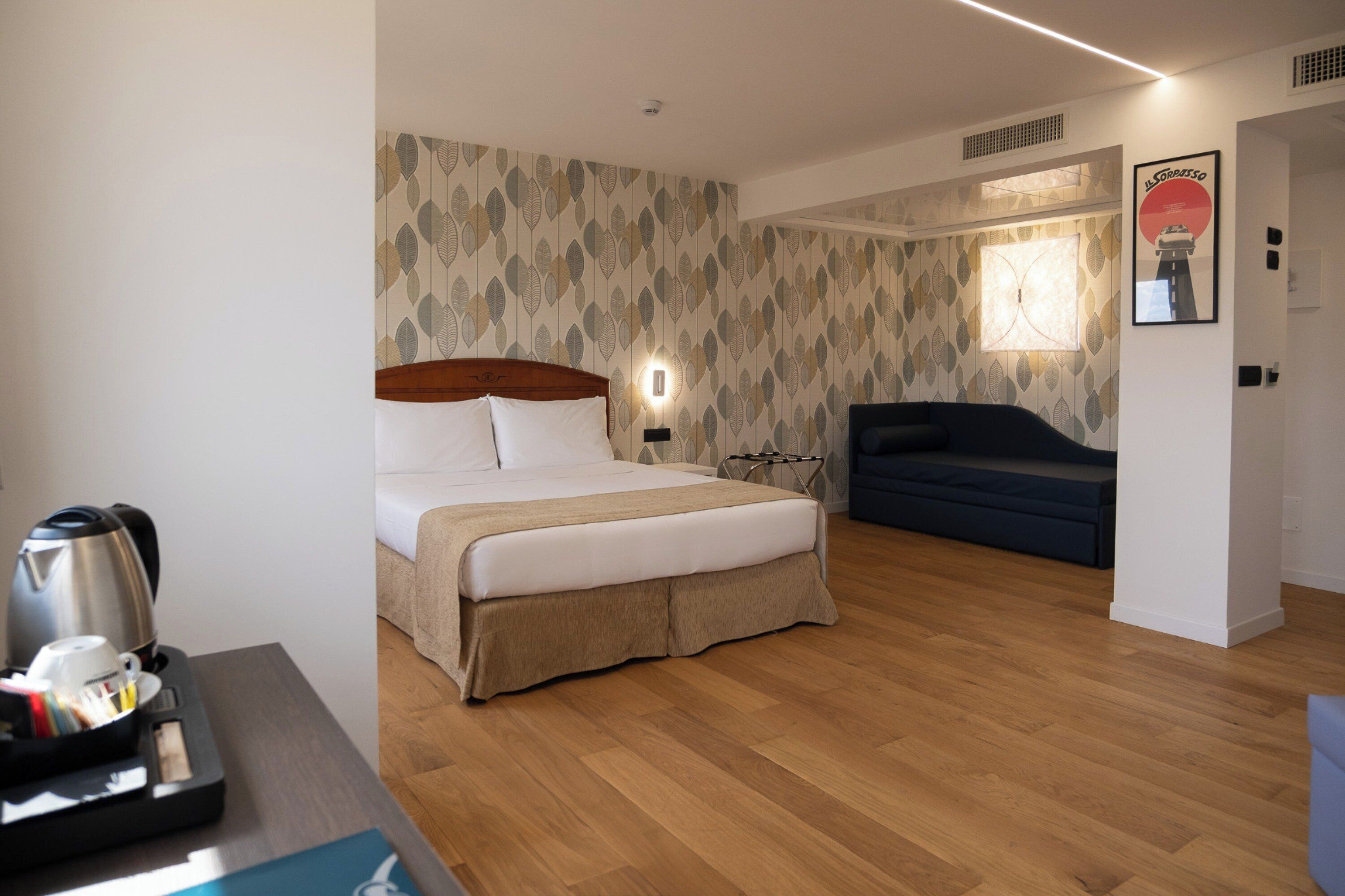 A hotel room with a bed, bedside table with a kettle and cups, a sofa, a wall with leaf-pattern wallpaper, and a poster.
