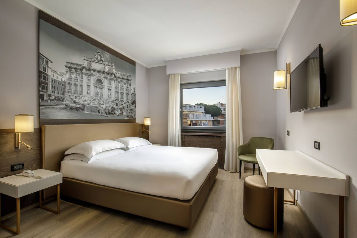 Habitación de hotel con una cama grande, mesitas de noche, un escritorio, una silla, un televisor y una ventana con cortinas.