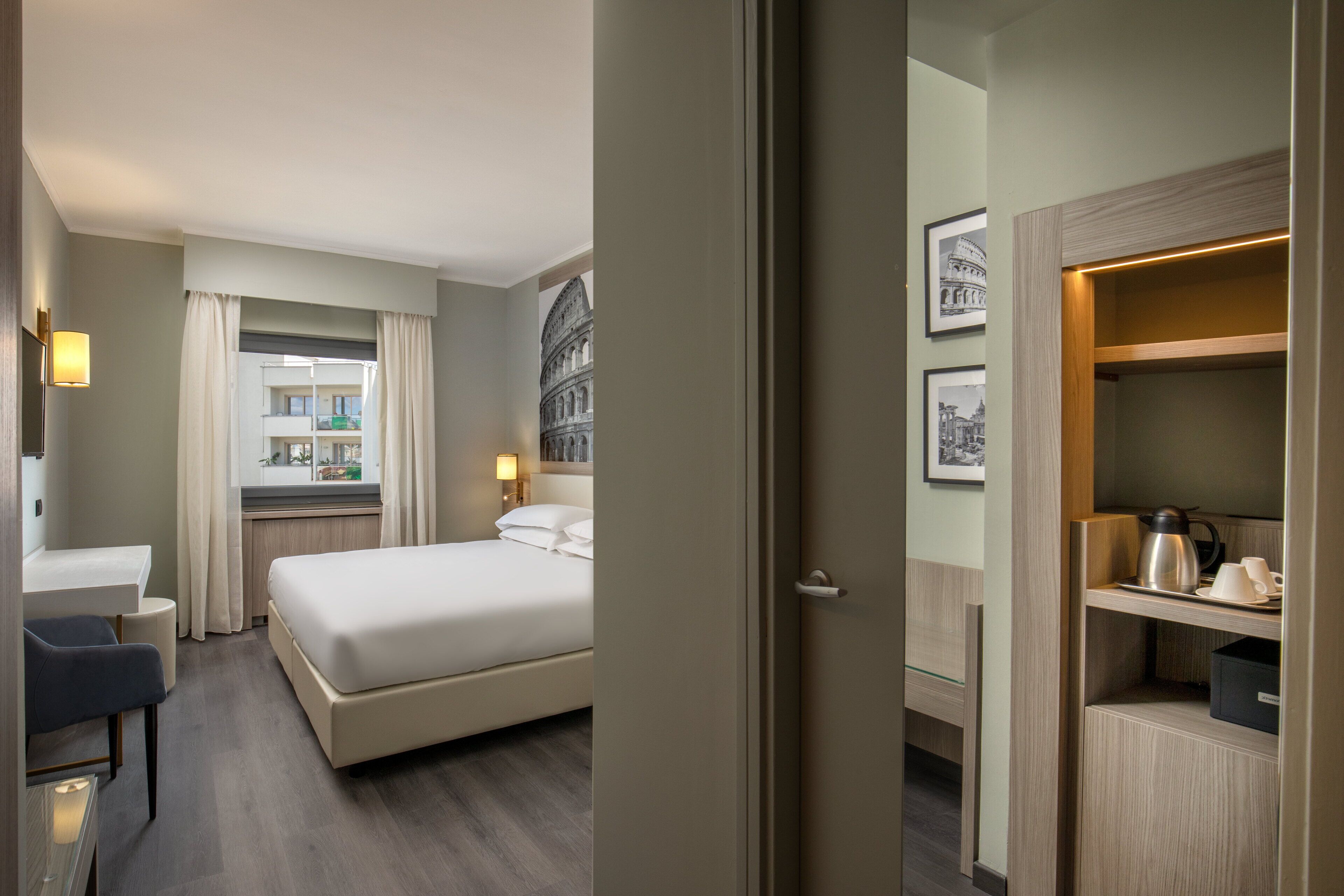 A hotel room with a bed, bedside table, lamp, and a view of a building through the window.