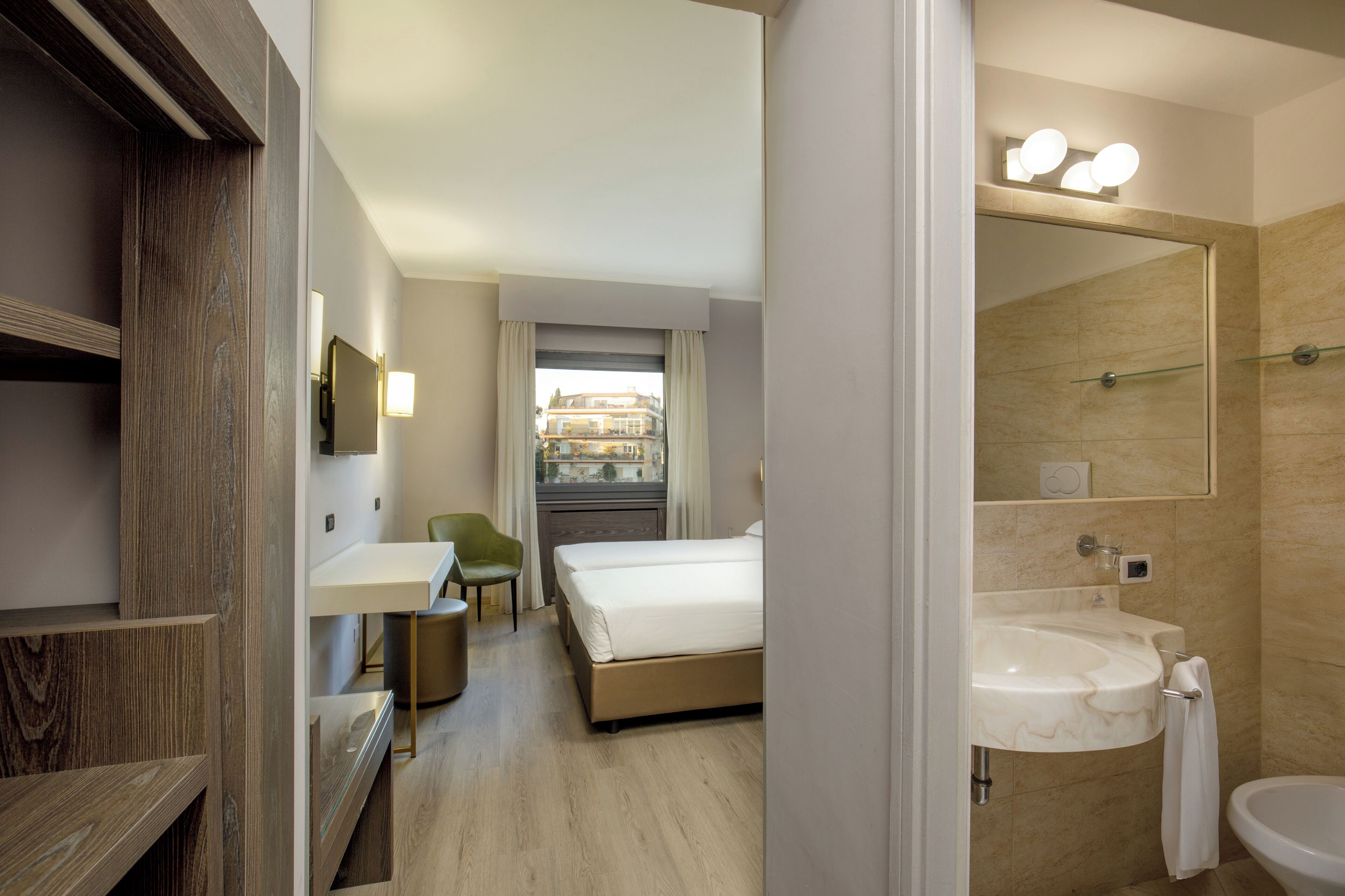 Une chambre d’hôtel comprenant un lit, un bureau, une chaise, une salle de bain avec un lavabo et un miroir, et donnant sur les bâtiments environnants.