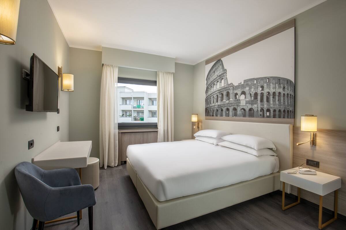 Habitación de hotel con una cama, un escritorio, una silla, un televisor y una pared decorada con una imagen del Coliseo.