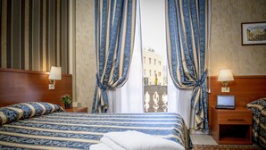 Une chambre d’hôtel avec un grand lit, deux tables de chevet avec des lampes, un ordinateur portable sur l’une des tables et une vue sur un immeuble à travers des rideaux transparents.