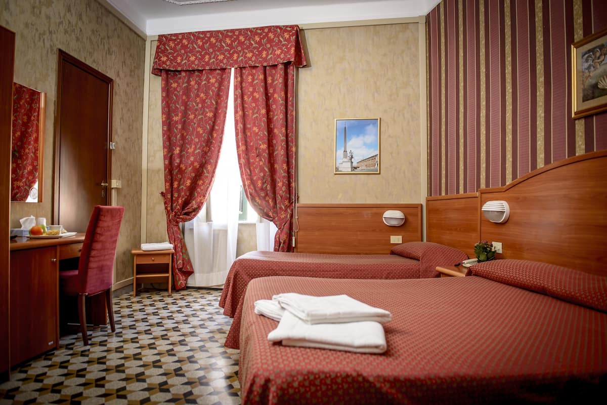A hotel room with two beds, a desk with a chair, a small table, and a window with curtains.
