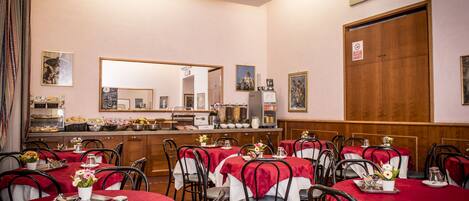 A dining area with tables covered in red tablecloths, white chairs, and a kitchen in the background.