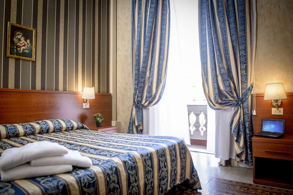 A hotel room with a bed, two bedside lamps, a laptop on a desk, and curtains.