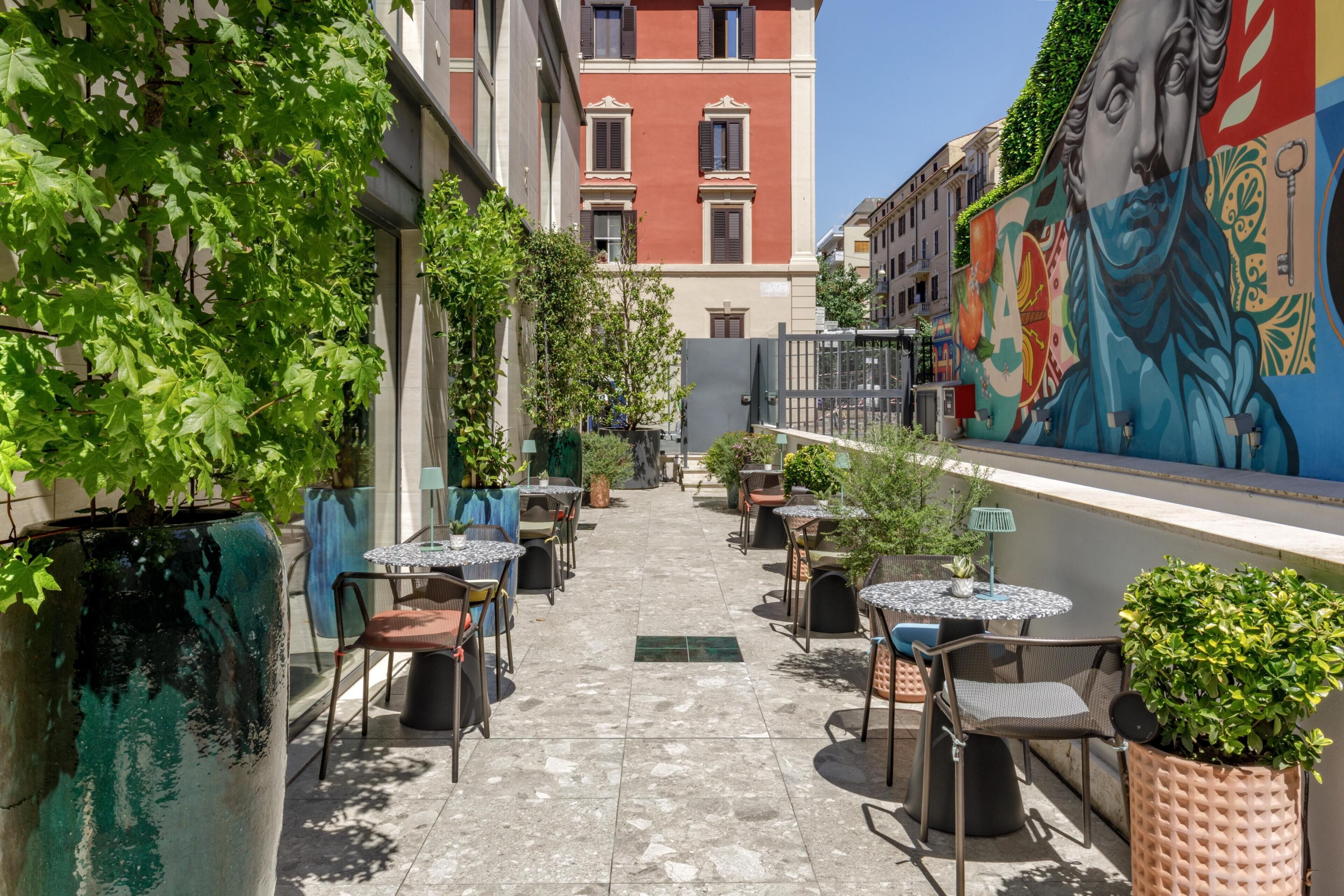 Un área de café al aire libre con mesas y sillas, rodeada de edificios y un mural.