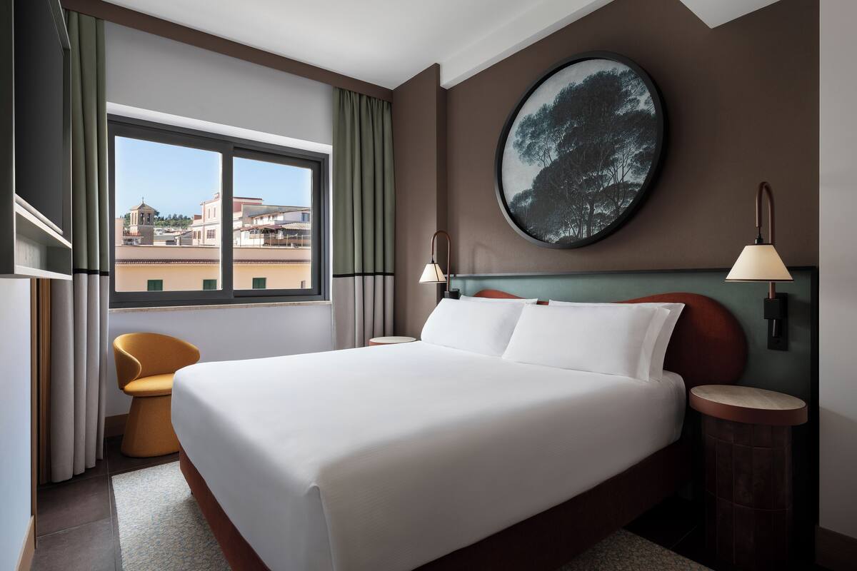 A hotel room with a large bed, a round painting on the wall, two bedside lamps, a view of buildings through the window, and a small round table.