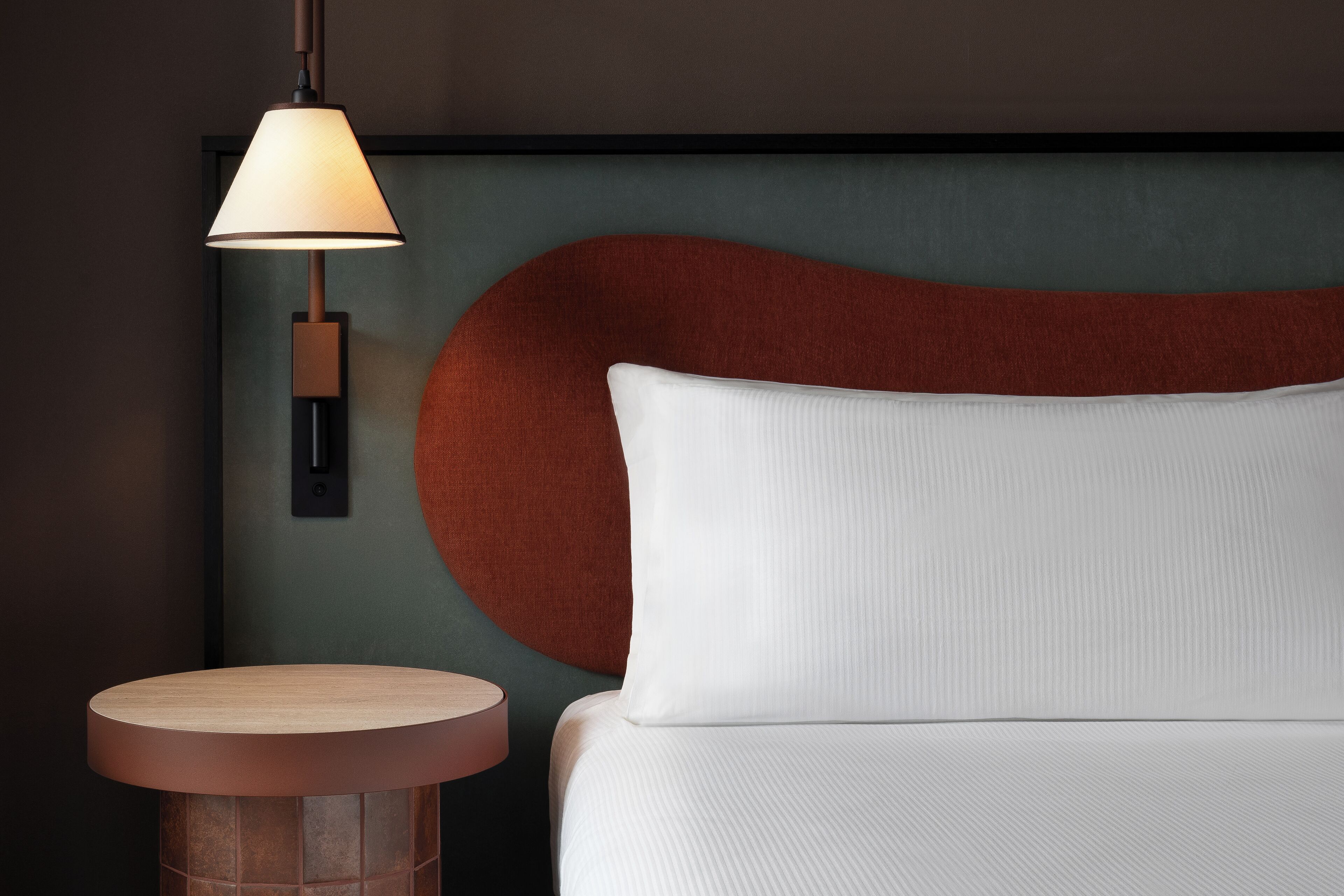 Una moderna camera d'hotel con un tavolino rotondo in legno, un letto bianco con cuscino decorativo e una lampada a parete.