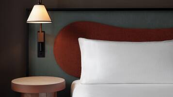 Una moderna camera d'hotel con un tavolino rotondo in legno, un letto bianco con cuscino decorativo e una lampada a parete.