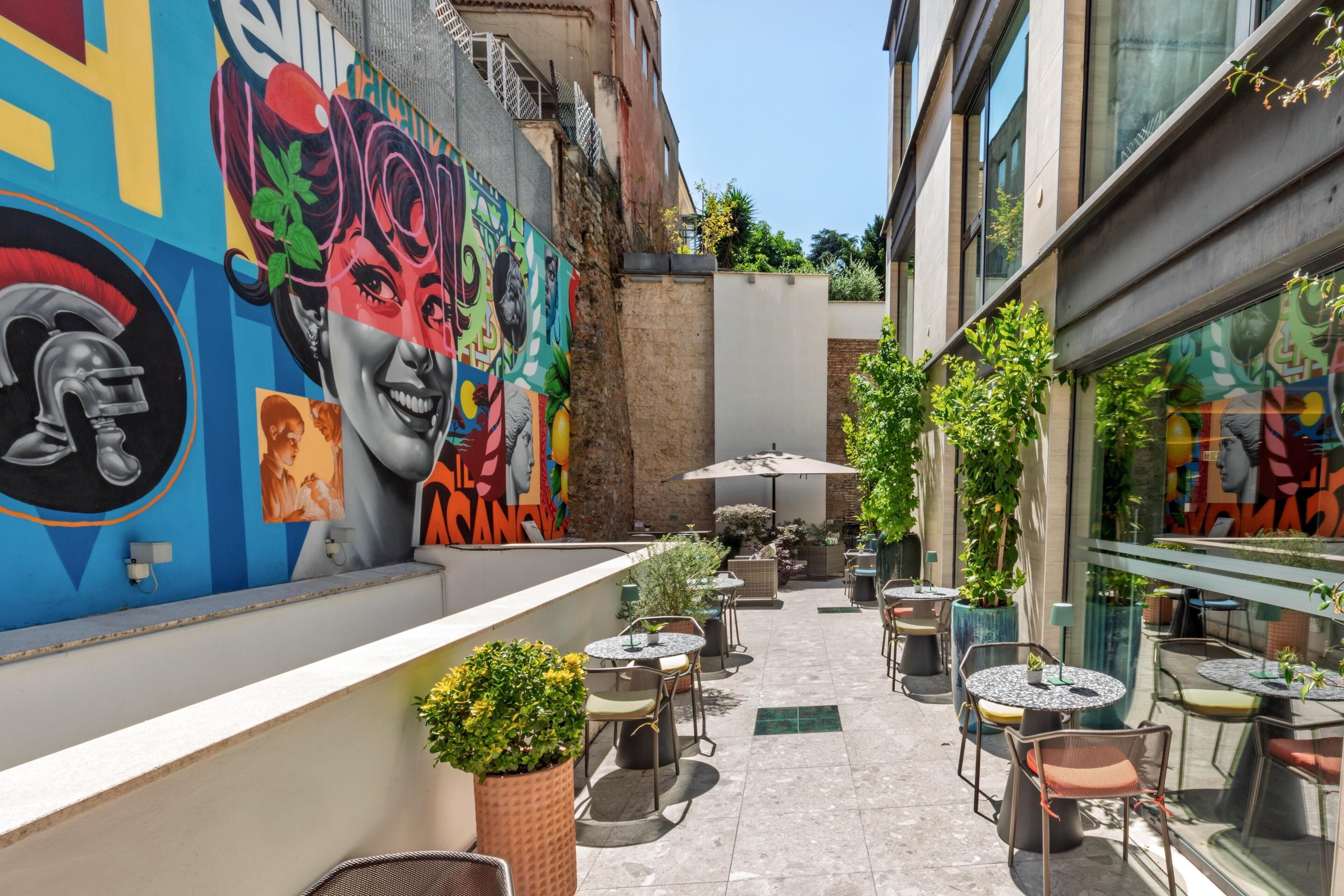 An outdoor café area with tables and chairs, a mural on the wall, and potted plants.