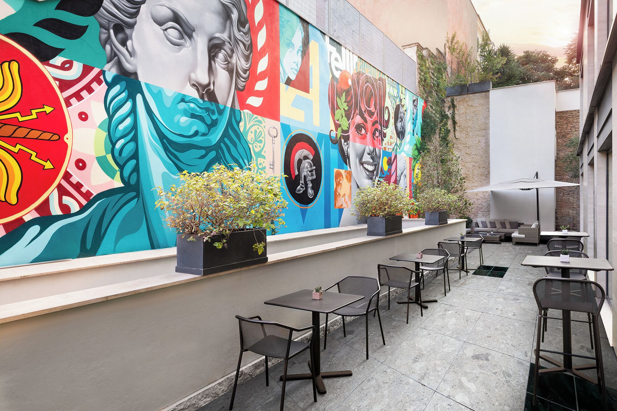 An outdoor patio with multiple tables and chairs, a mural with various artistic elements, and potted plants.
