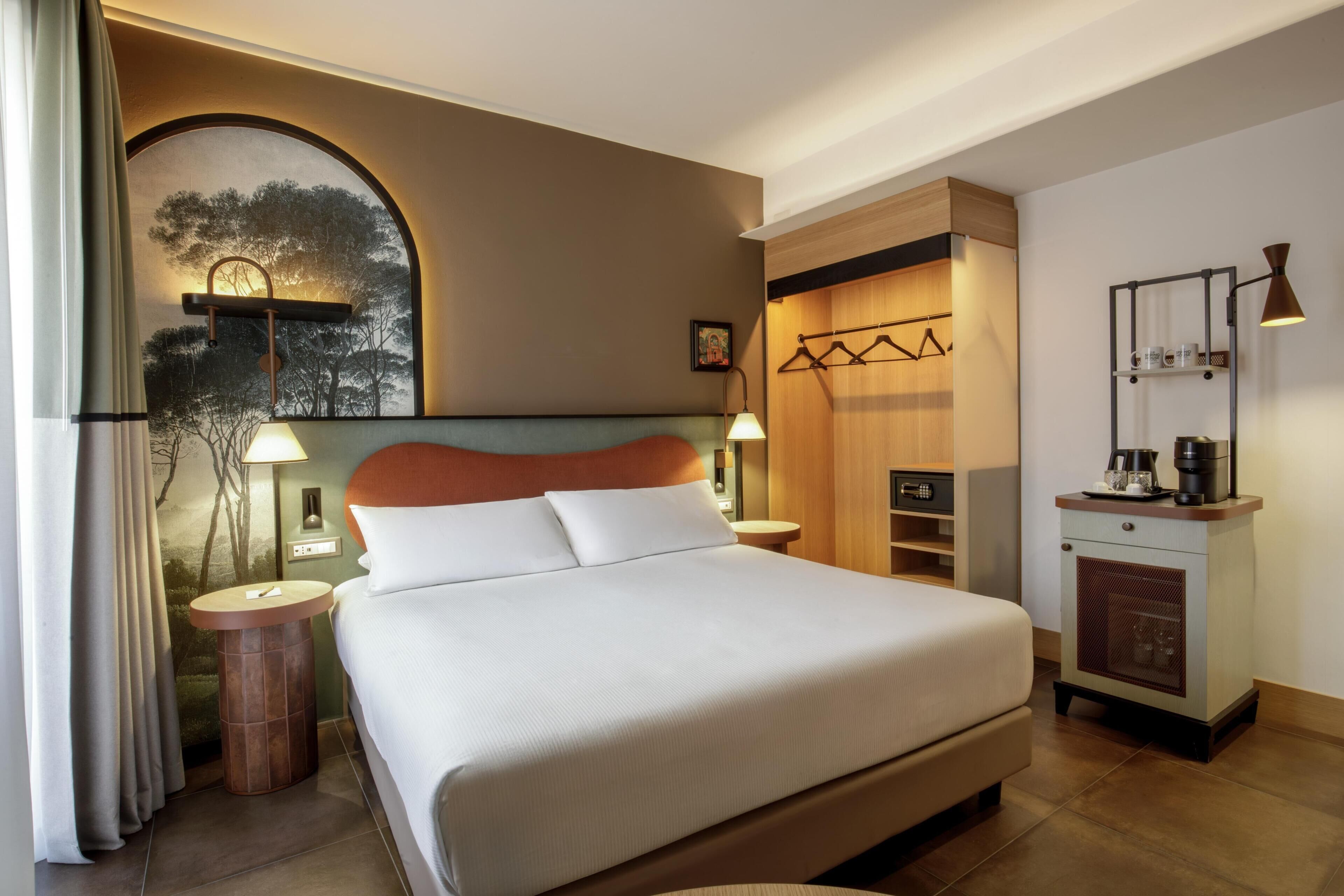 A hotel room with a large bed, bedside tables, a wardrobe, a small desk with a lamp, and a view of trees through a window.