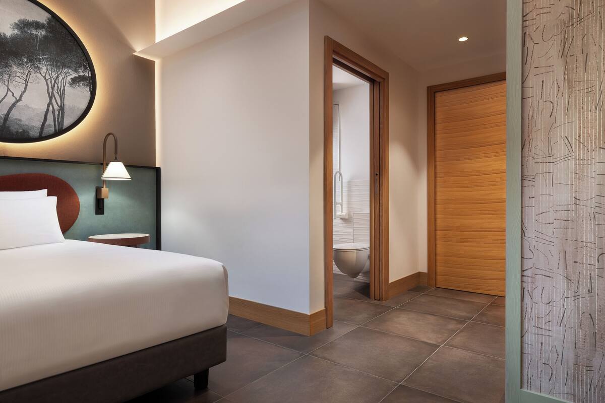 A modern hotel room with a large bed, a round wall-mounted light, and a view of the bathroom through an open door.