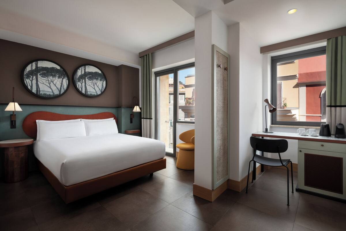 A hotel room with a bed, a desk, a chair, two wall art pieces, and a window with a view of buildings.