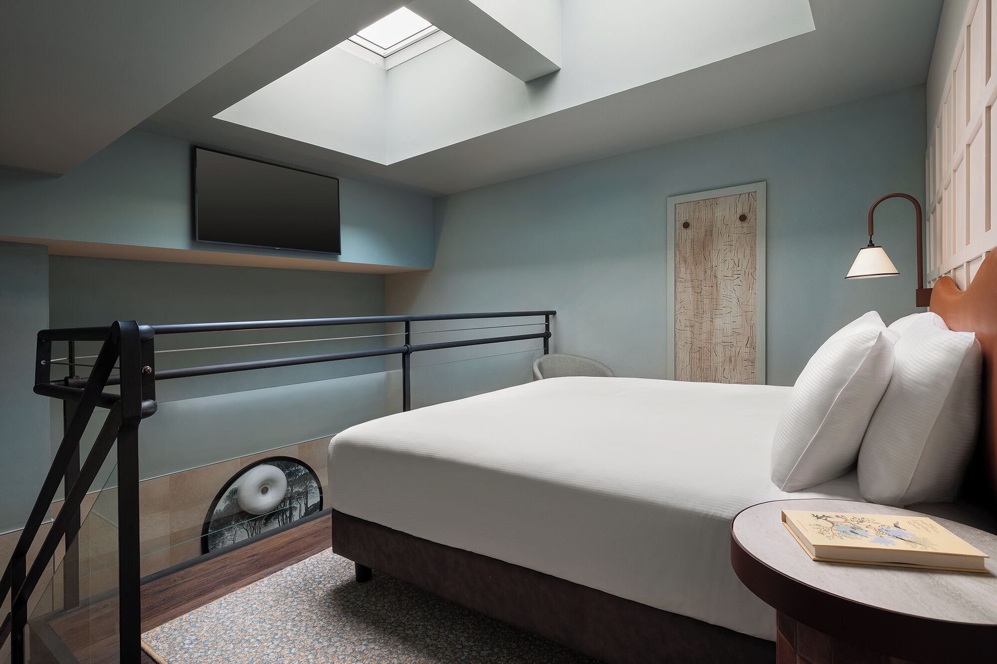 A modern bedroom with a skylight, a bed, a bedside table with a book, and a wall-mounted TV.