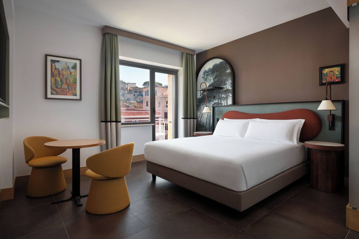 A hotel room with a large bed, two chairs, a small table, and a view of buildings through the window.