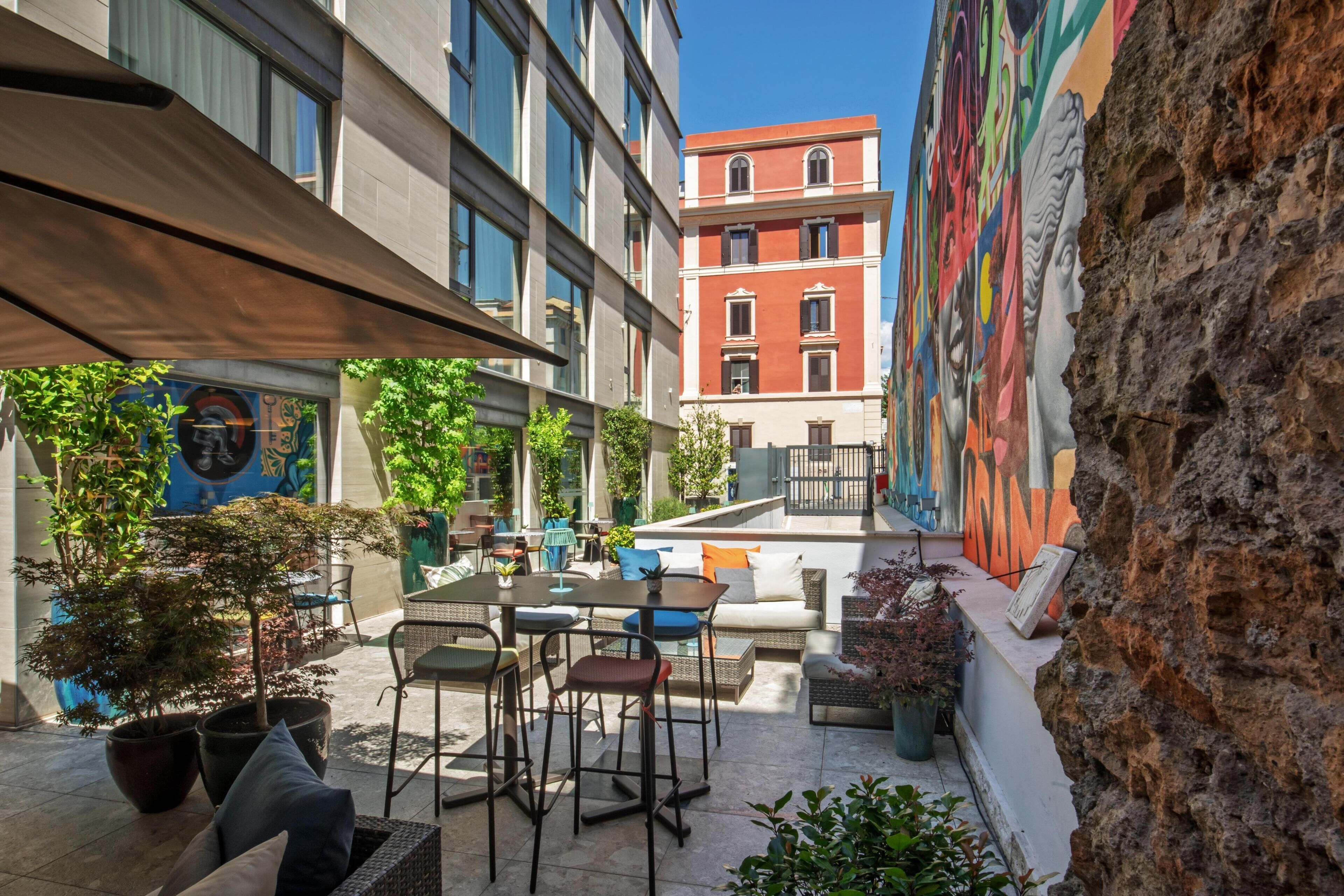 Un área de estar al aire libre con mesas y sillas, rodeada de macetas con plantas y un mural en la pared.