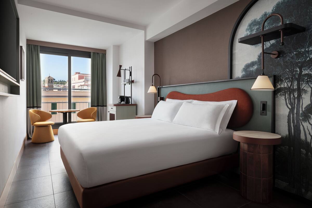 A modern hotel room with a large bed, a desk, two chairs, and a view of the cityscape.
