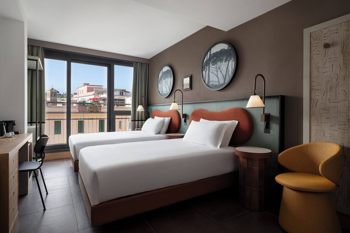 A hotel room with two beds, a desk, and a chair. There are large windows with a view of buildings outside.