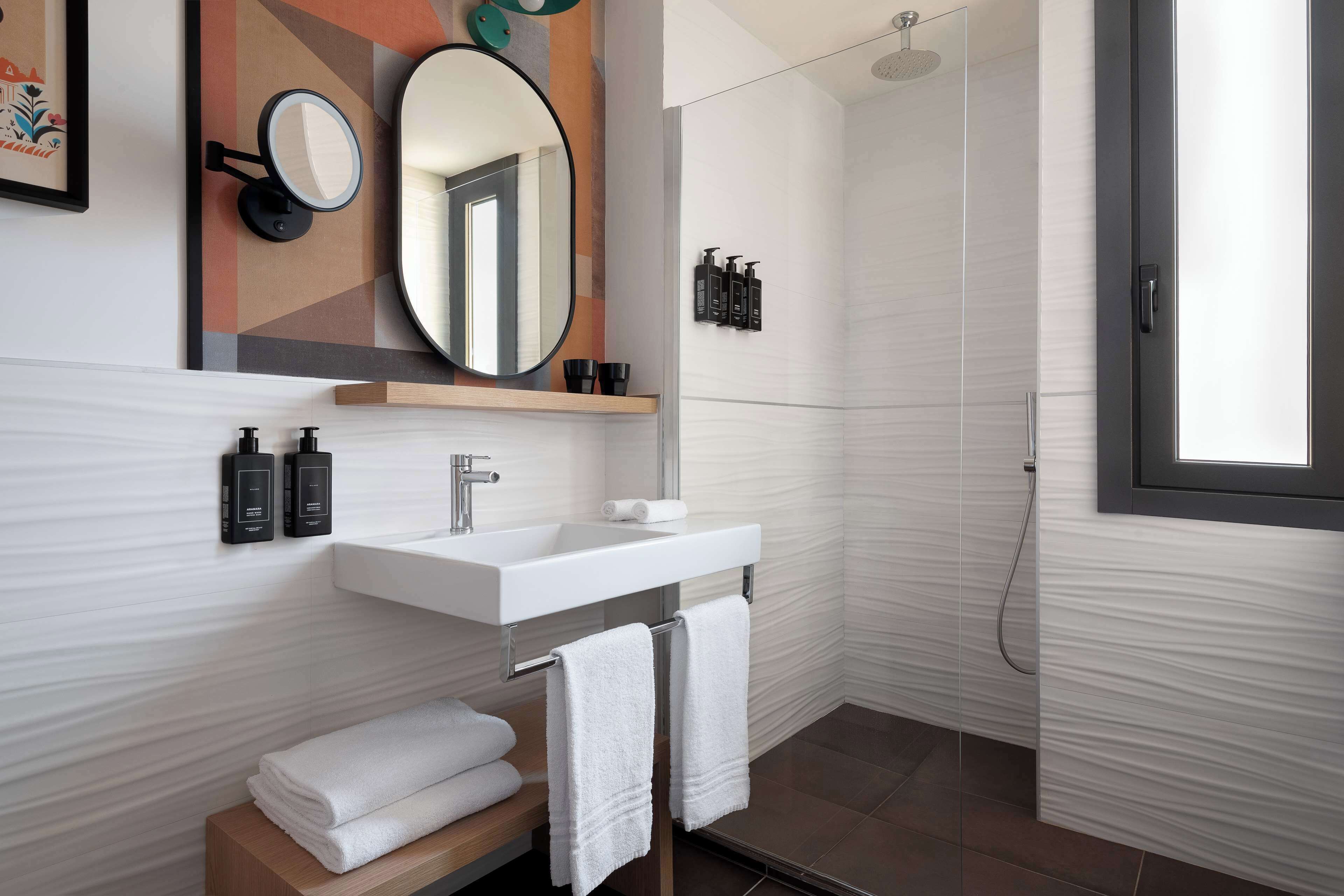 Un bagno moderno con un lavandino bianco, uno specchio e una doccia. Asciugamani e articoli da toilette sono presenti sul lavandino.