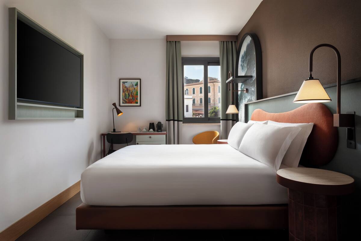 A modern hotel room with a large bed, a desk, a chair, a lamp, and a view of buildings through the window.