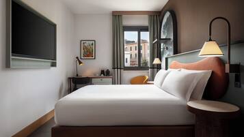 Habitación de hotel moderna con una cama grande, un escritorio, una silla, una lámpara y vistas a edificios a través de la ventana.