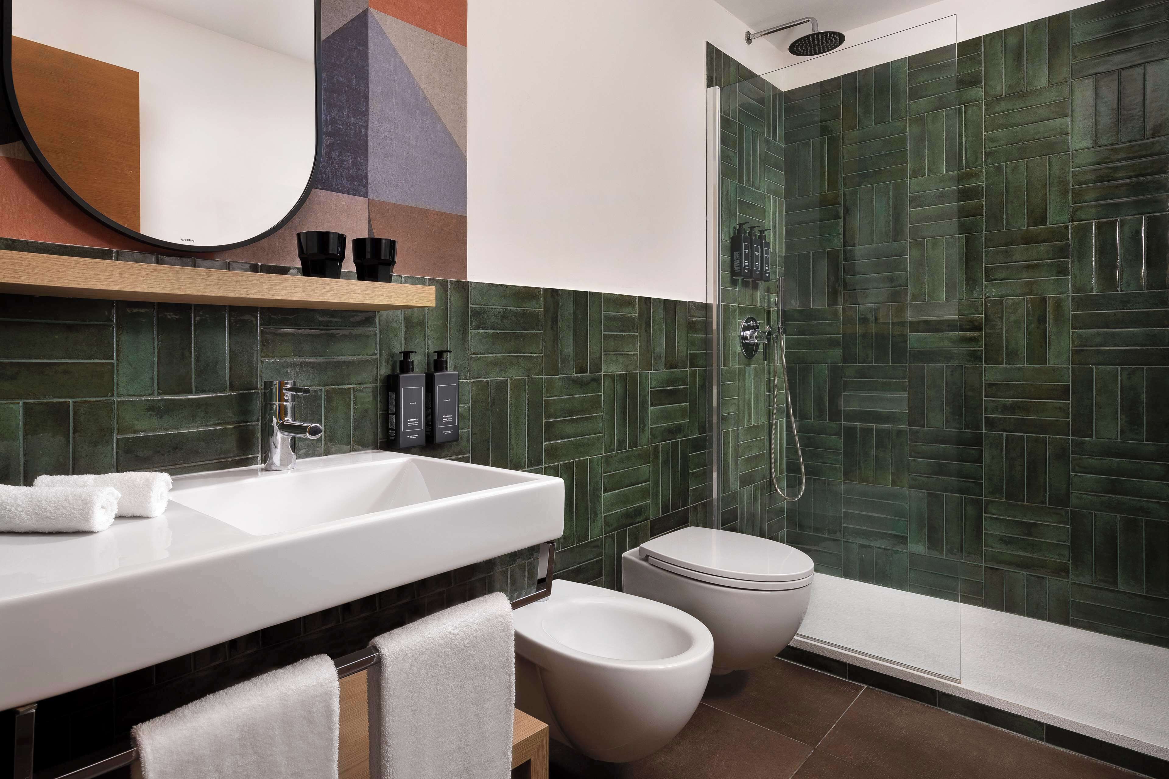 A modern bathroom with a white sink, two toilets, a shower area with green tiles, and a mirror with a geometric design above the sink.