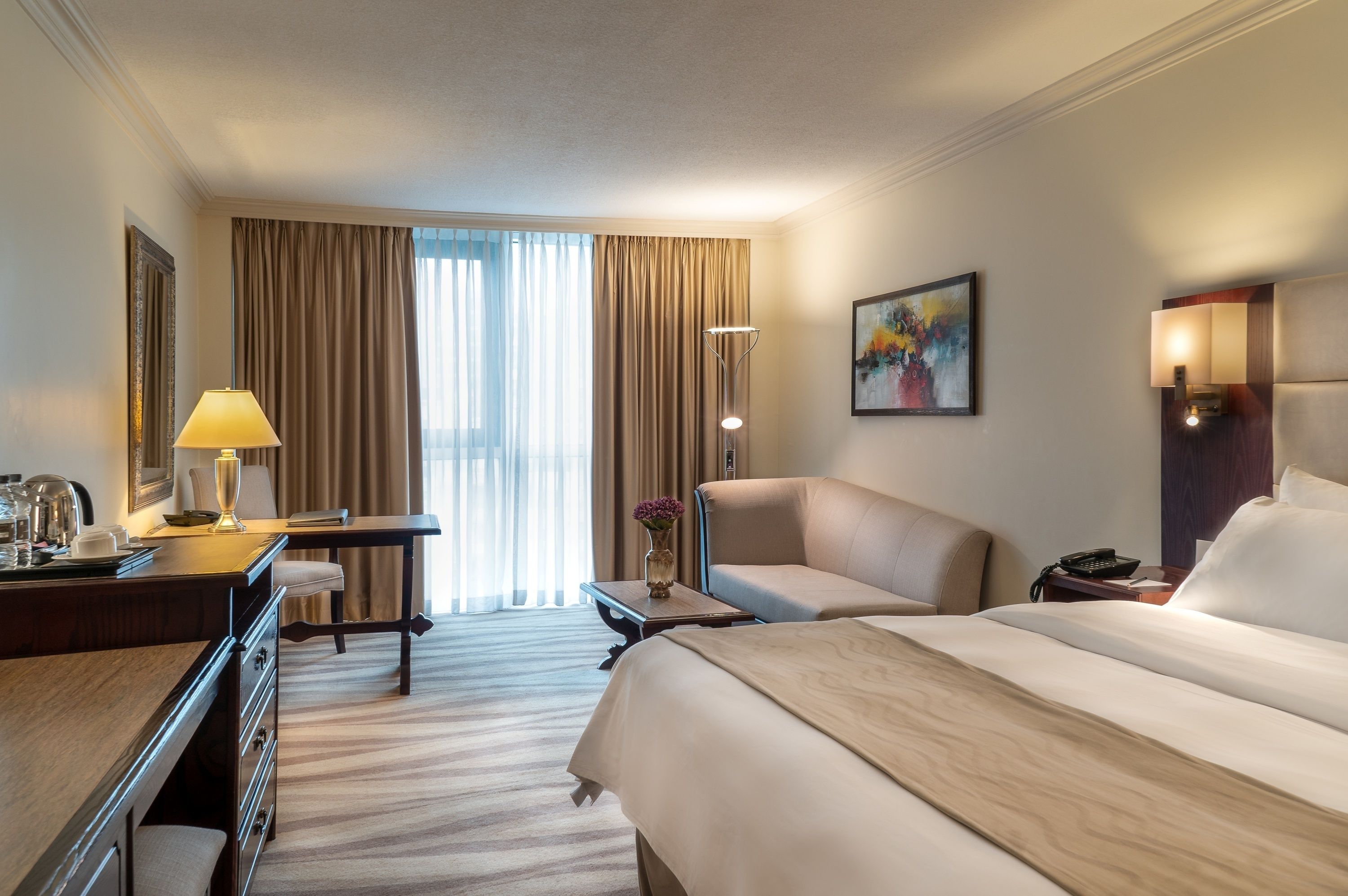 A hotel room with a large bed, a desk with a lamp, a chair, a small table with a vase, and a painting on the wall.