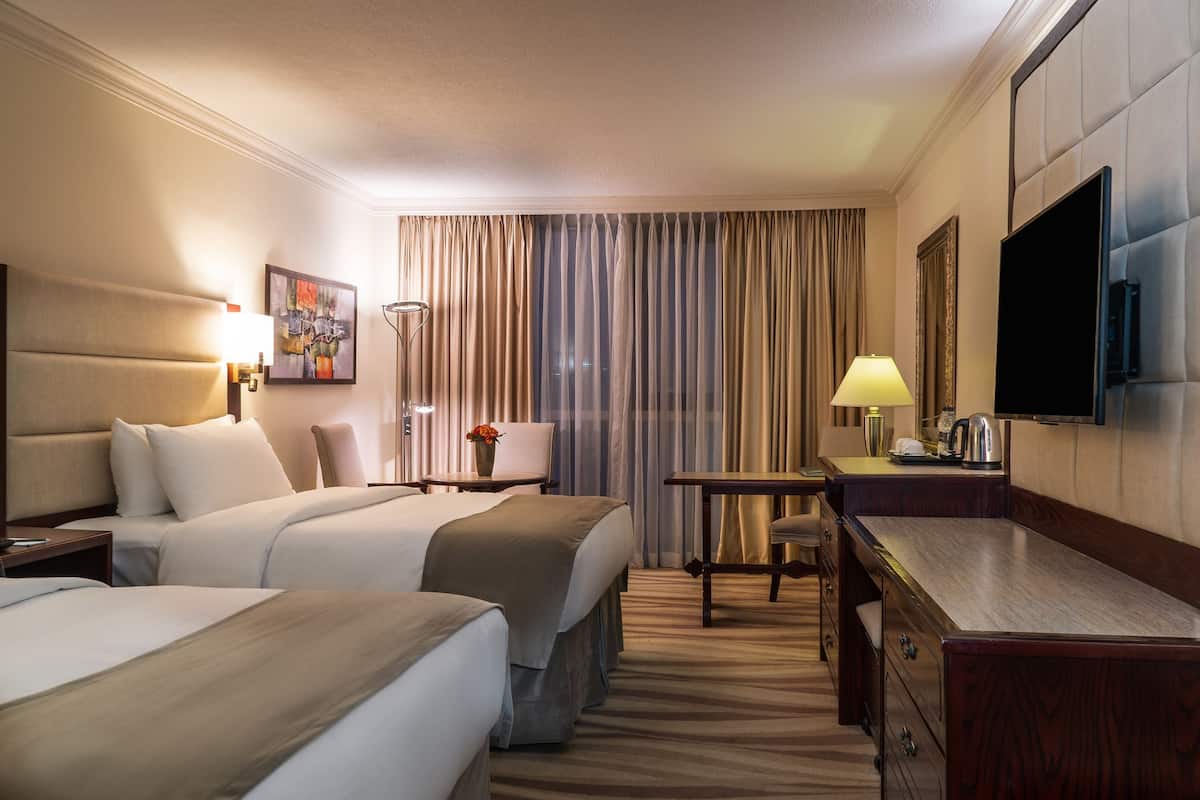 A hotel room with two beds, a desk, a chair, a television, and a large window with curtains.