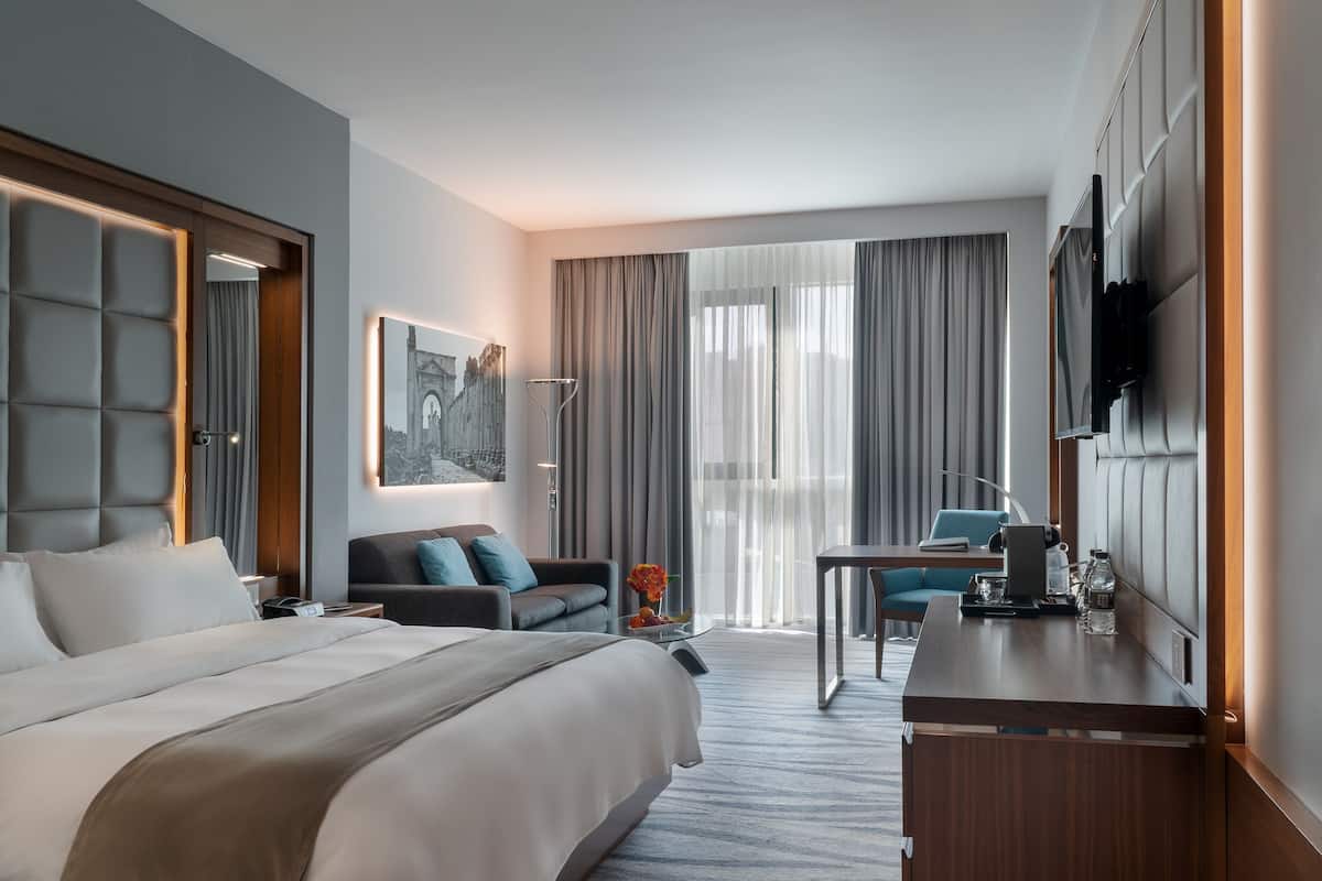 A modern hotel room with a large bed, a desk, a chair, a TV, and a window with curtains.