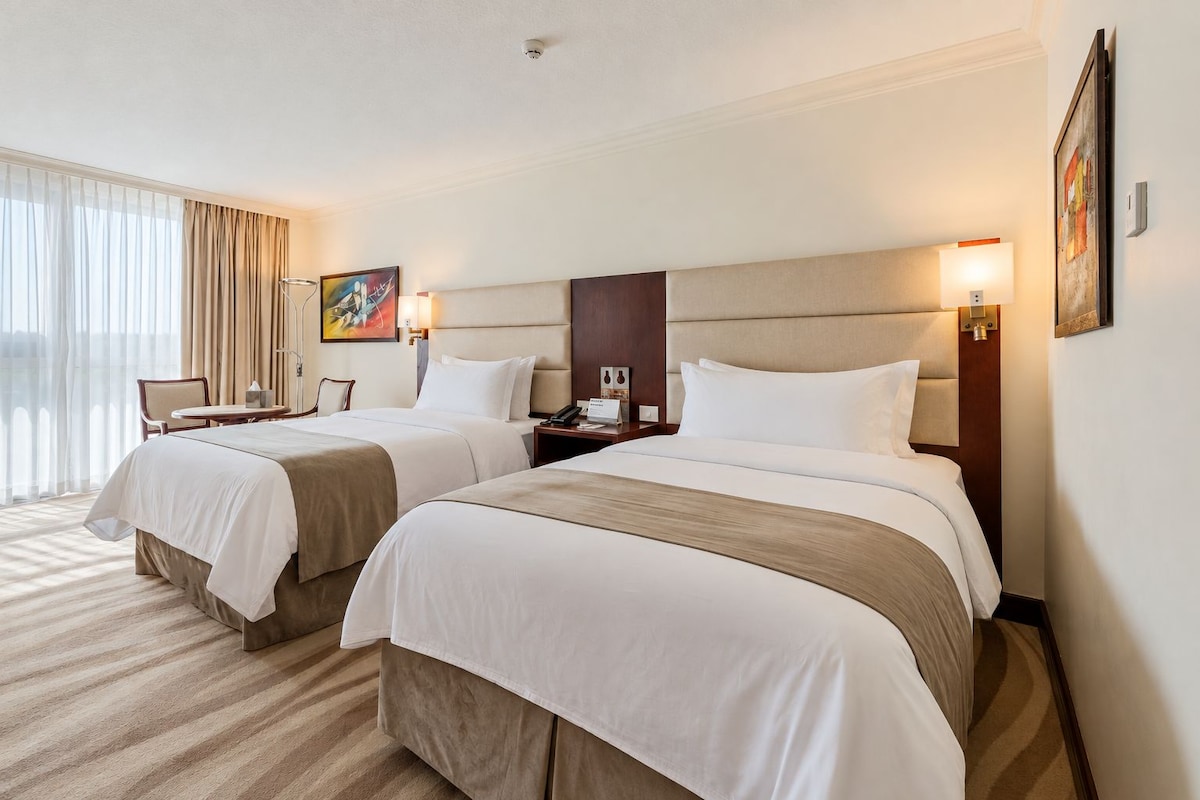 A hotel room with two beds, a desk, a chair, and a large window with curtains.