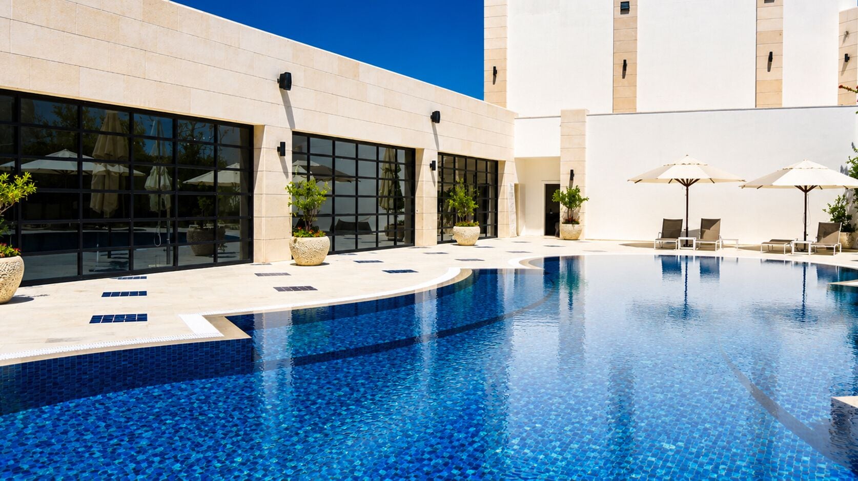 Une piscine entourée de chaises longues et de parasols, le tout bordé d’un bâtiment doté de grandes fenêtres et agrémenté de plantes en pot.