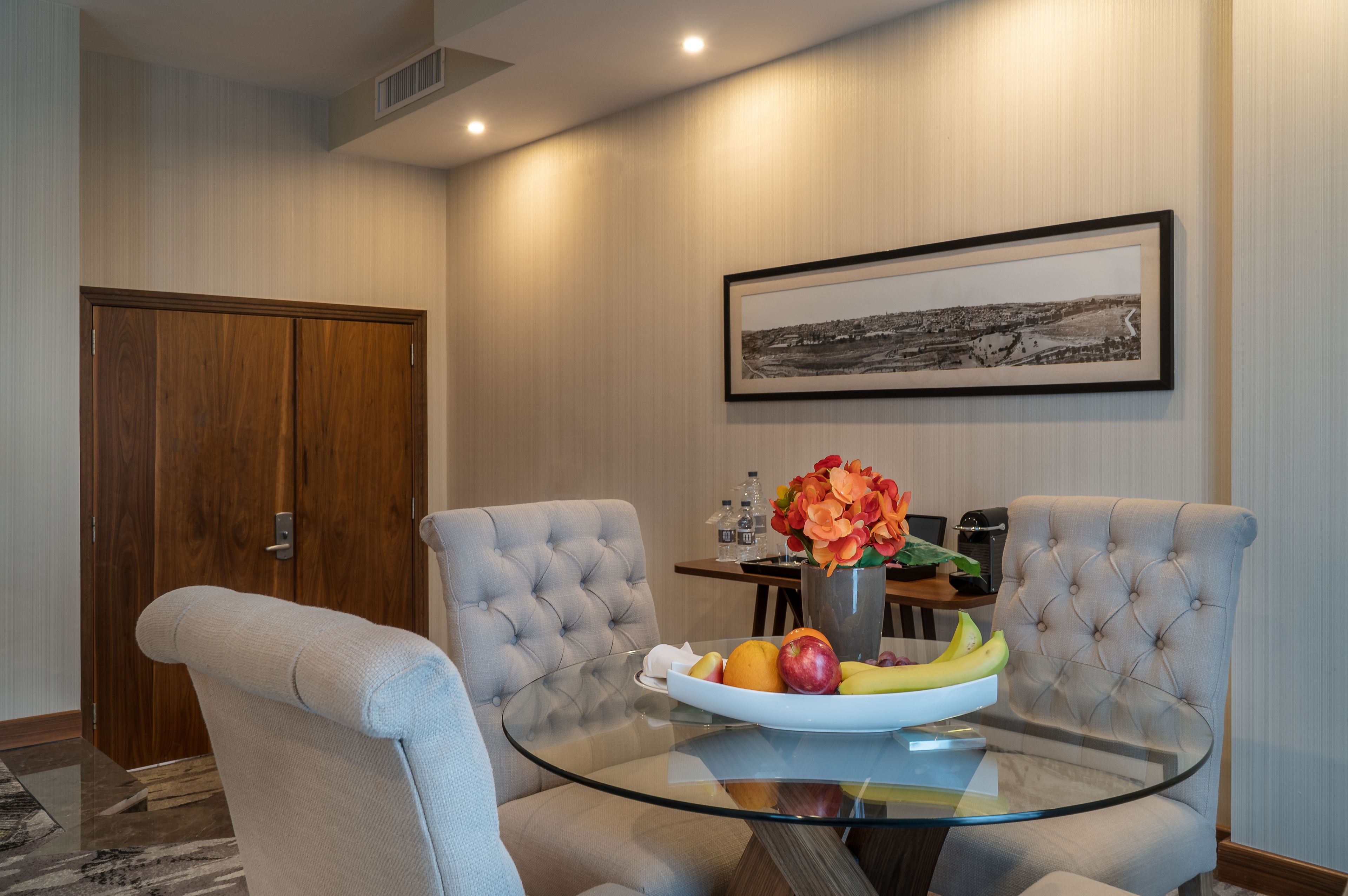 A dining area with a glass table, two upholstered chairs, a framed picture on the wall, and a vase with flowers.