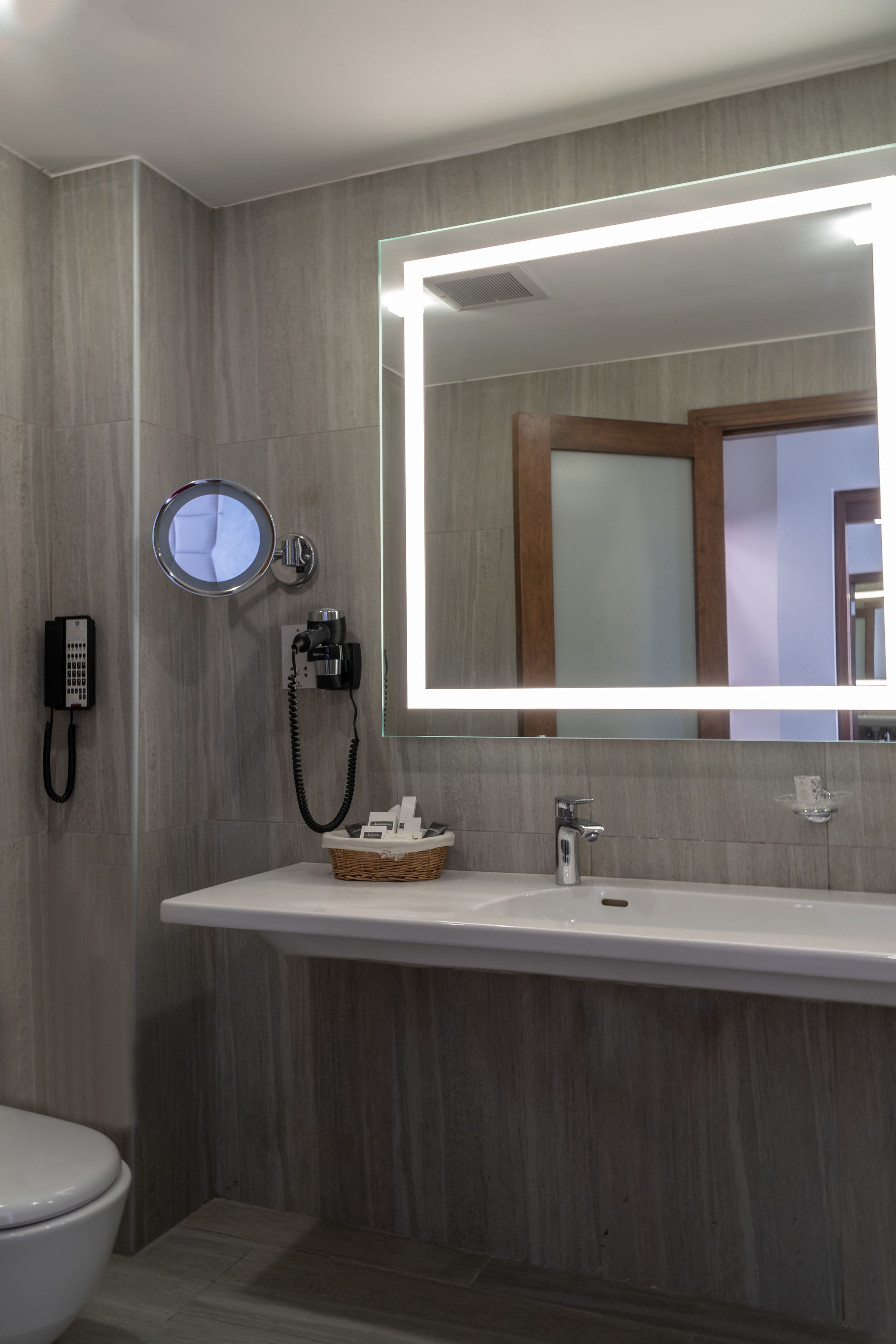 A modern bathroom with a large mirror, a round vanity mirror, a built-in heater, a telephone, and a basket of toiletries.