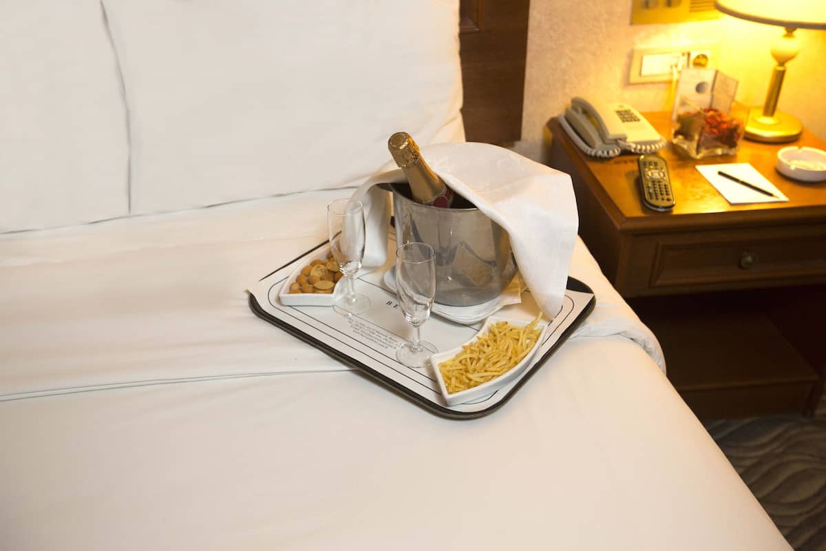 Habitación de hotel con cama, mesita de noche con lámpara, teléfono y bandeja con champán, copas y aperitivos.