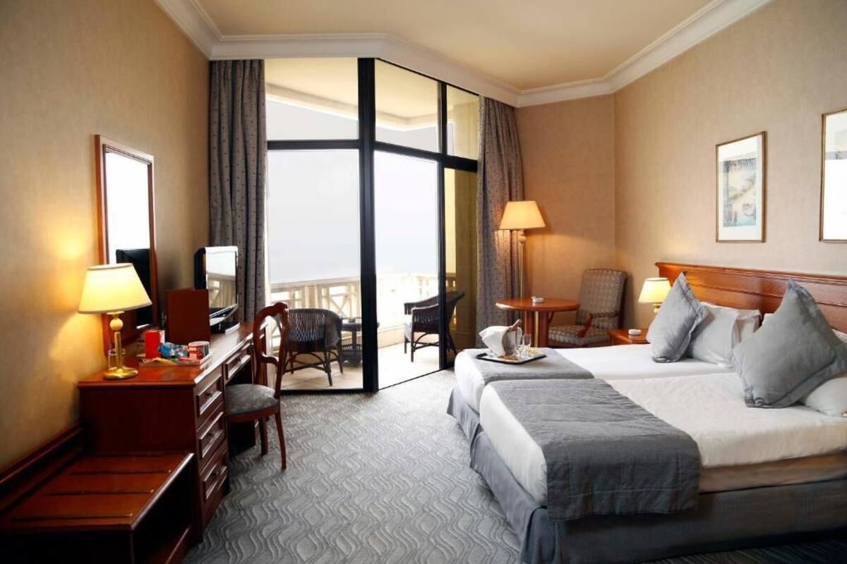 Habitación de hotel con una cama grande, un escritorio con lámpara, una silla, una mesita con lámpara y vistas a un área exterior a través de un ventanal.