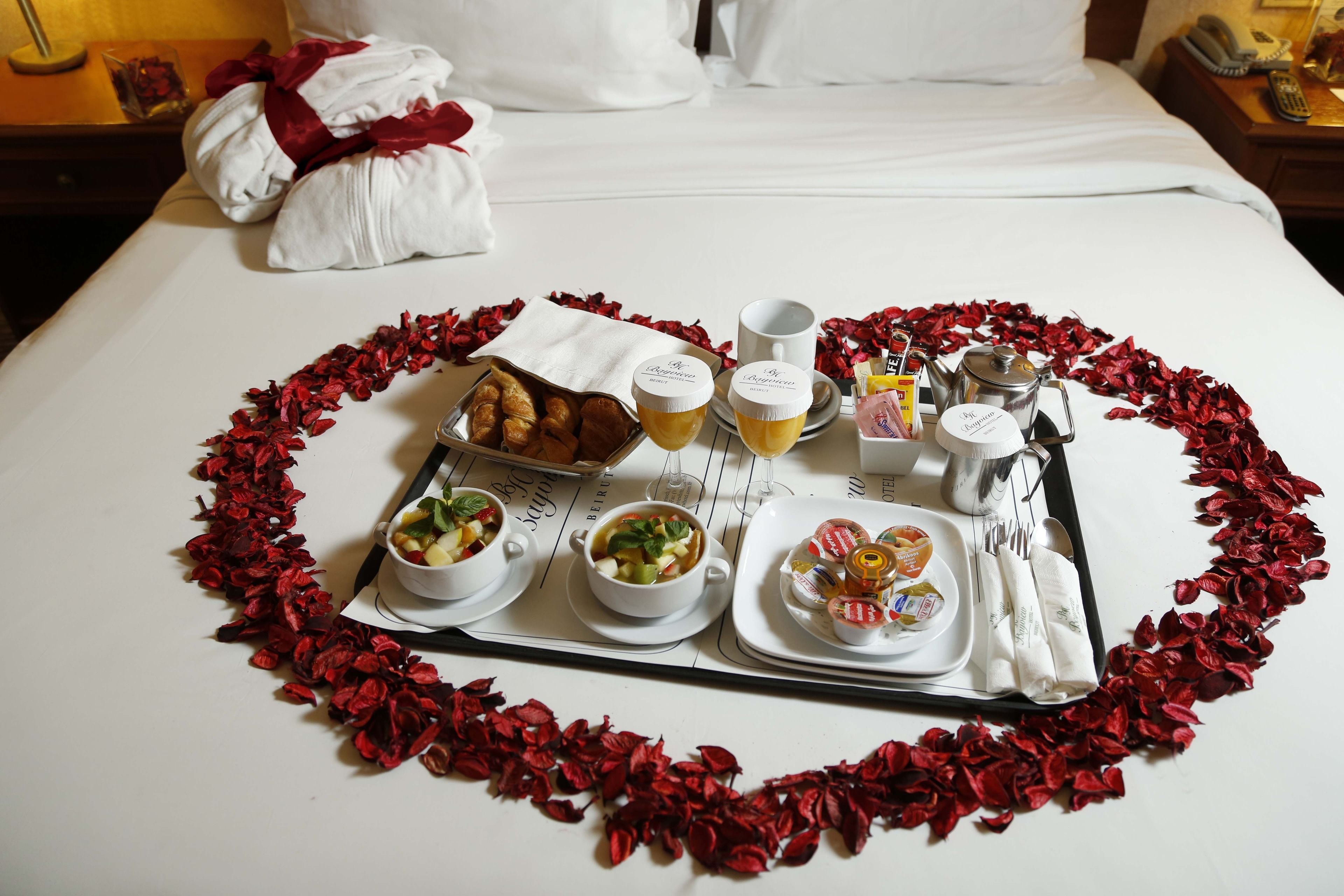 A neatly arranged breakfast tray on a bed with a heart-shaped arrangement of rose petals, including croissants, fruit, and beverages.