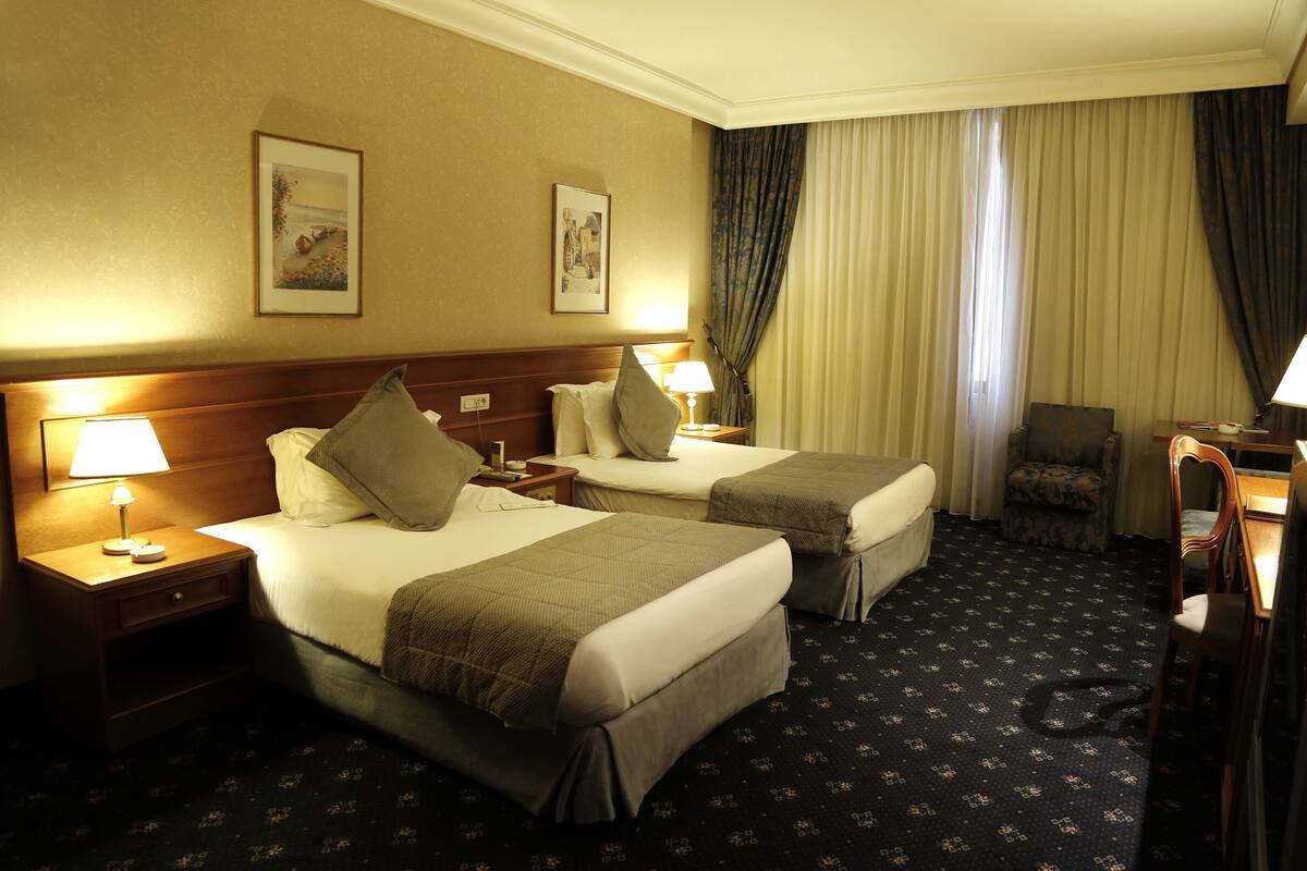 Habitación de hotel con dos camas, una mesita de noche con lámpara, una silla y un escritorio con lámpara.