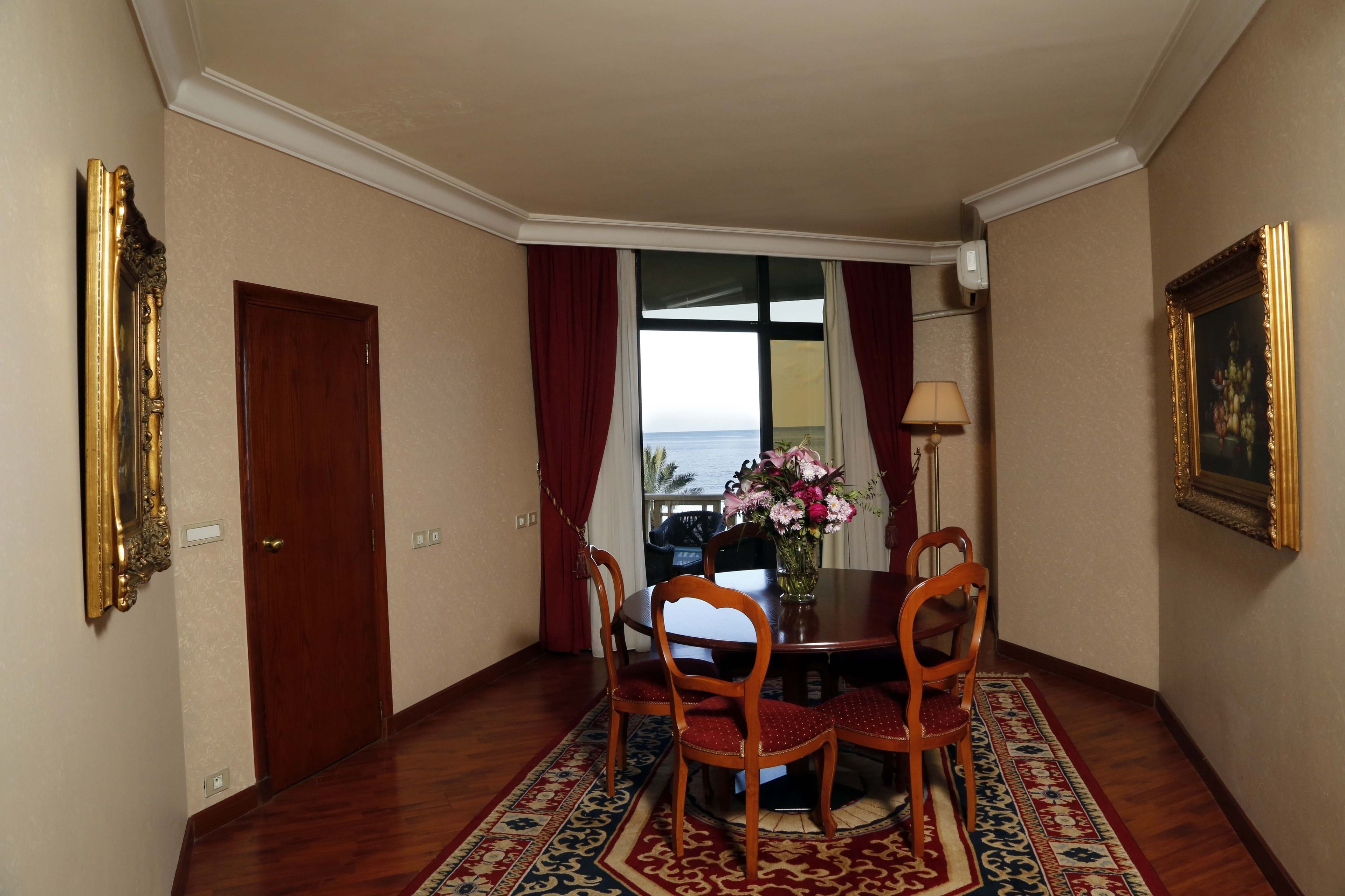 A dining area with a table and chairs, a vase with flowers, and a view of the sea through a window.