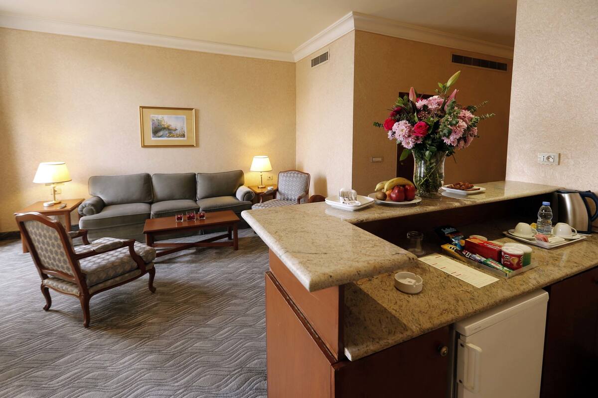 Habitación de hotel con sofá, sillón, mesa de centro y pequeña cocina equipada con mostrador y electrodomésticos.