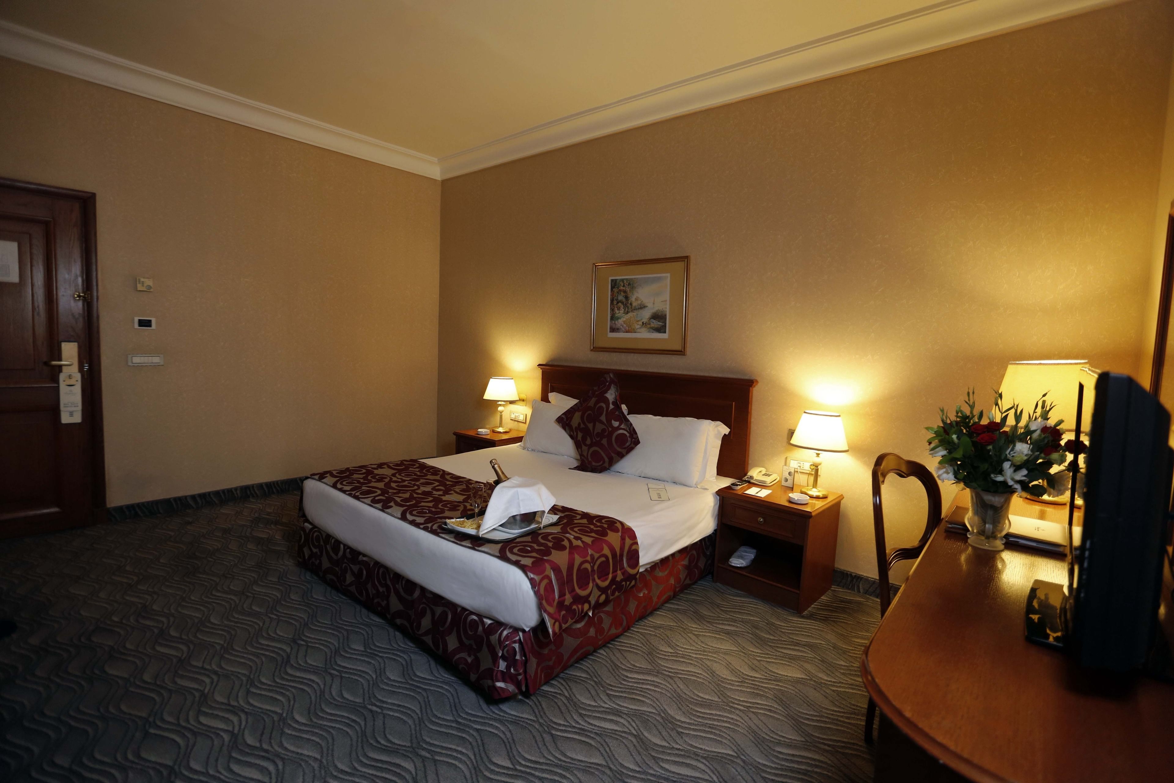 Habitación de hotel con una cama grande, dos mesitas de noche con lámparas, una silla de madera y un escritorio con un florero.