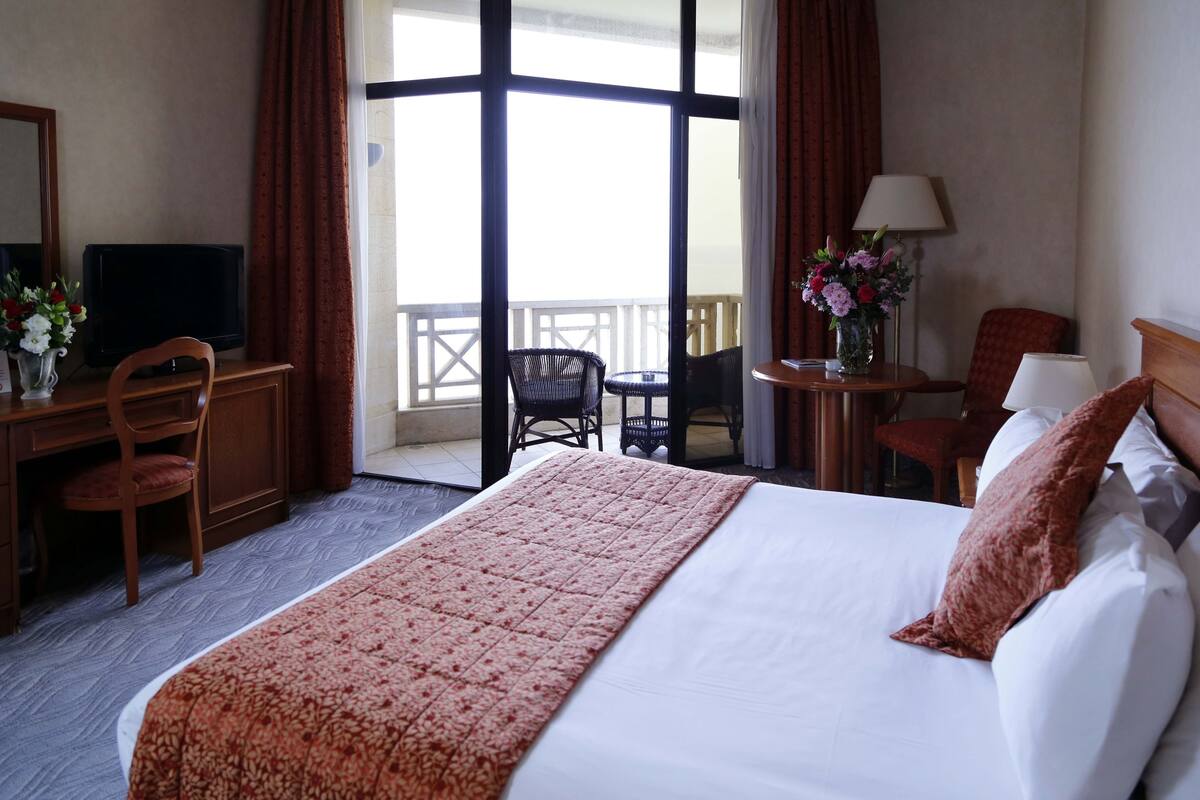 Habitación de hotel con cama, escritorio con televisor, silla, mesita de noche con lámpara y balcón con vistas.
