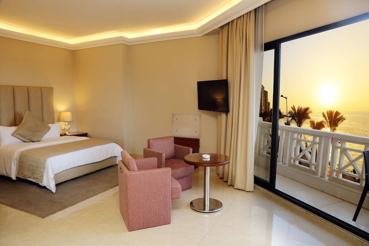 Habitación de hotel con una cama, dos sillones rosas, una mesita, un televisor y un balcón con vista al mar.