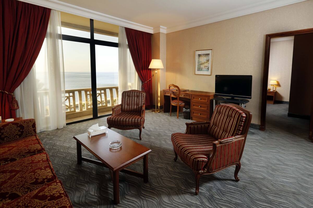 Habitación de hotel con un ventanal grande, un sofá, dos sillones a rayas, una mesa de centro de madera, un escritorio con un televisor y una lámpara.
