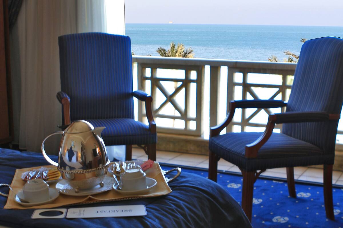 Habitación de hotel con cama, dos sillones, una bandeja con juego de té y vista al océano.