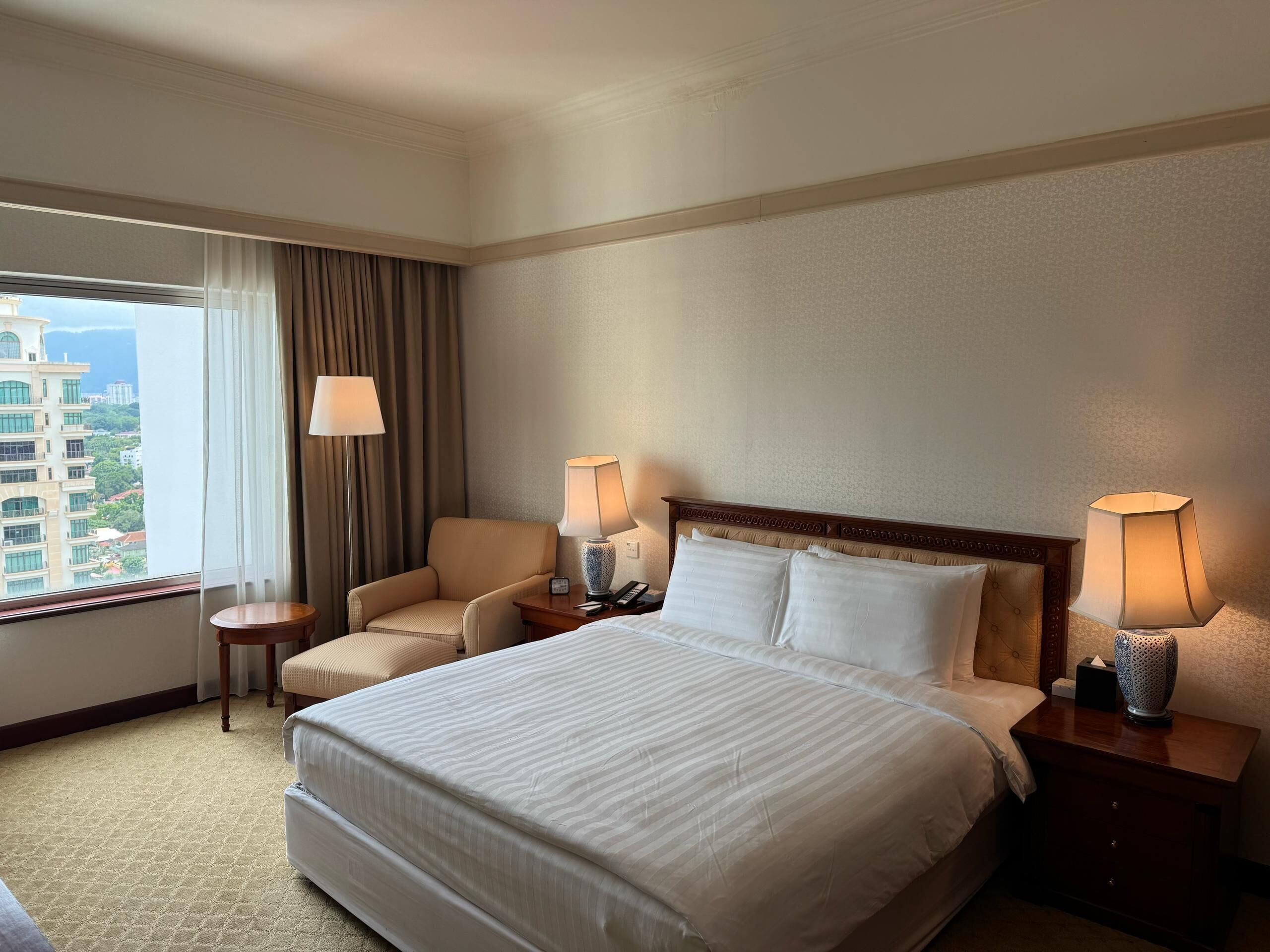 A hotel room with a large bed, two bedside tables with lamps, a chair, and a view of a cityscape through the window.