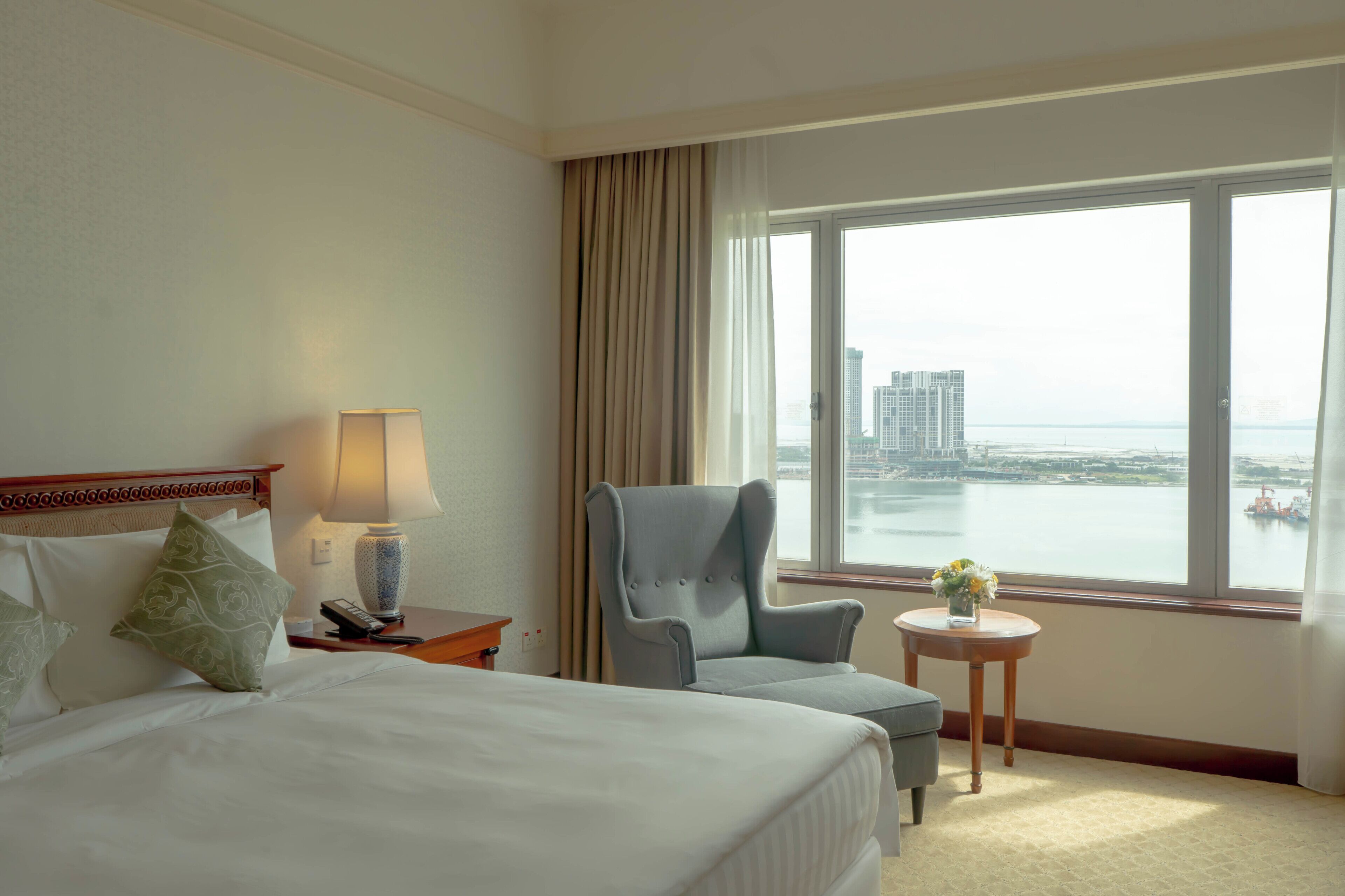 Ein Hotelzimmer mit Bett, einem Sessel, einem kleinen Tisch mit Vase und Blick auf eine Stadtlandschaft durch ein großes Fenster.
