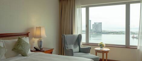 A hotel room with a bed, a chair, a small table with a vase, and a view of a cityscape through a large window.