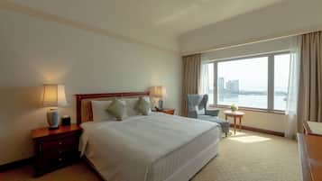 A hotel room with a large bed, two bedside tables with lamps, a chair, a small table with a vase, and a large window with a view of buildings and water.